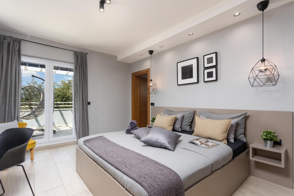 A modern bedroom features a spacious bed adorned with neutral linens and decorative cushions. A small nightstand displays a potted plant, while framed artwork is showcased on the wall. Natural light floods through double patio doors leading to a balcony.