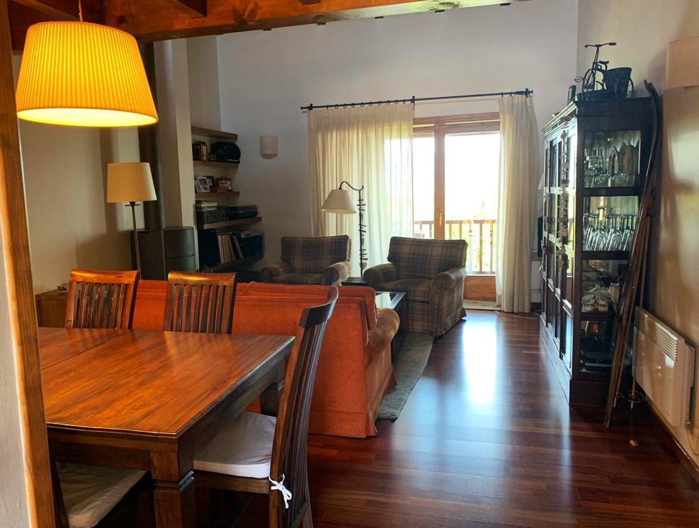 A comfortable living area is furnished with two plaid chairs and a brown sofa, positioned near a large window draped with sheer curtains. A wooden dining table with several chairs is located in the foreground, complemented by warm lighting from a pendant lamp.