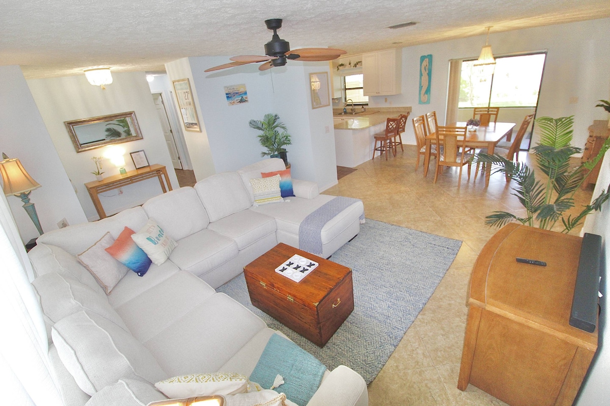 A spacious living area features a comfortable sectional couch and a wooden coffee table, creating a welcoming space. A ceiling fan hangs above, providing airflow. In the background, a dining area with a light wood table and chairs is visible, along with an open kitchen that allows for easy interaction.