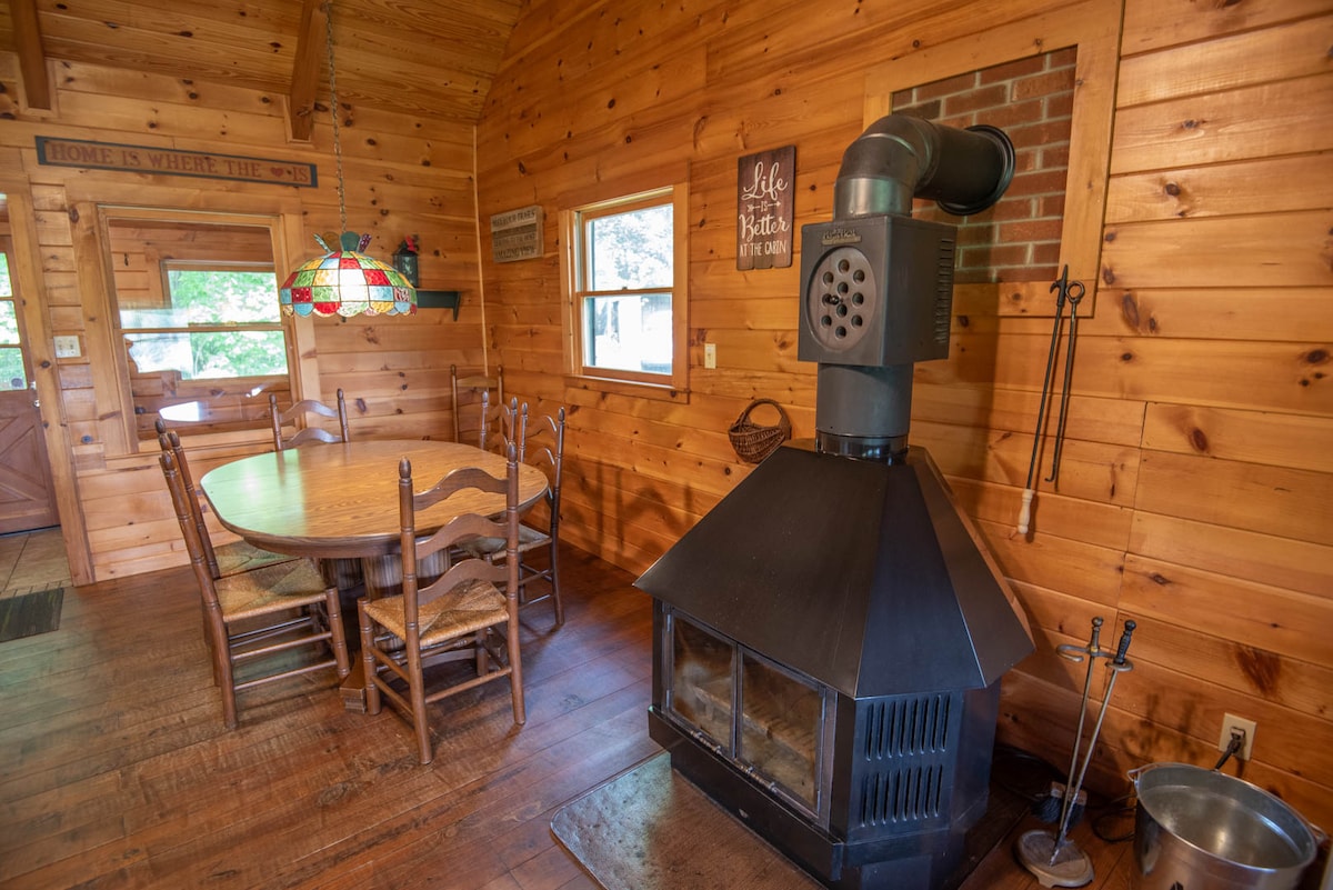 A cozy dining area features a wooden table surrounded by six rustic chairs. A wood stove is positioned against a wooden wall, with its chimney leading to the ceiling. Natural light streams through nearby windows, illuminating the warm wood paneling.