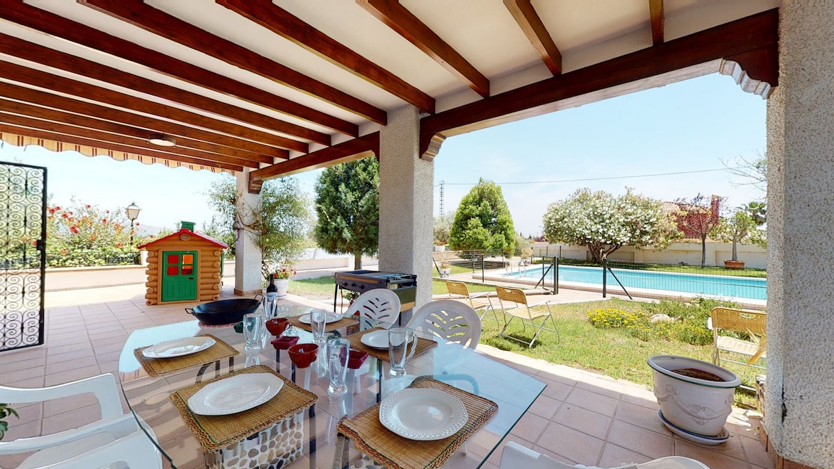 An outdoor dining area features a glass table set with plates and cutlery. The space is shaded by a wooden roof, providing a view of a private fenced pool and landscaped garden, complemented by trees and flowering plants.