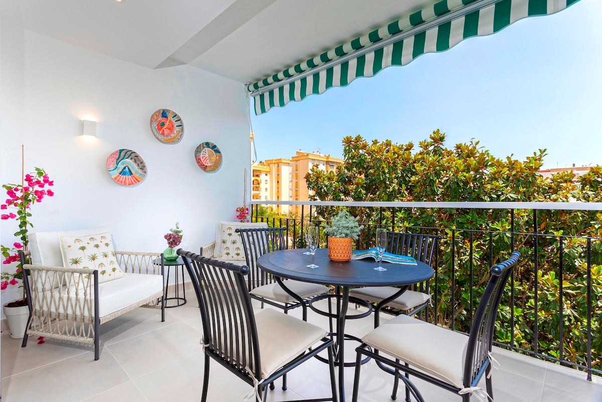 A private terrace is shown, featuring a round dining table surrounded by black chairs and comfortable armchairs. Colorful wall decor and potted plants add vibrancy. A green striped awning provides shade, with views of greenery beyond, enhancing the outdoor seating area.
