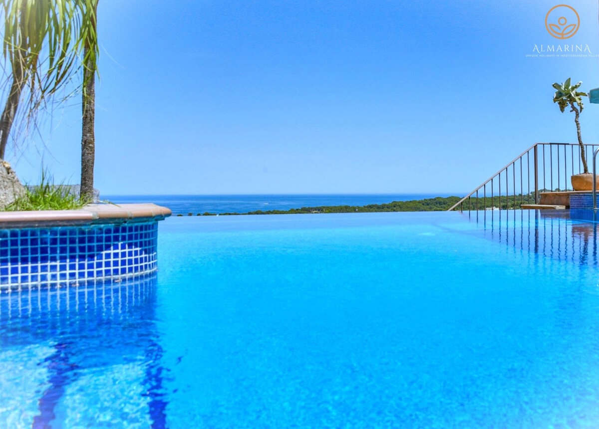 An inviting infinity pool stretches towards panoramic views of the Mediterranean Sea. The clear blue water beautifully reflects the bright sky, while surrounding palm trees add a touch of greenery. A gentle slope leads to the area beyond the pool, enhancing the scenic landscape.
