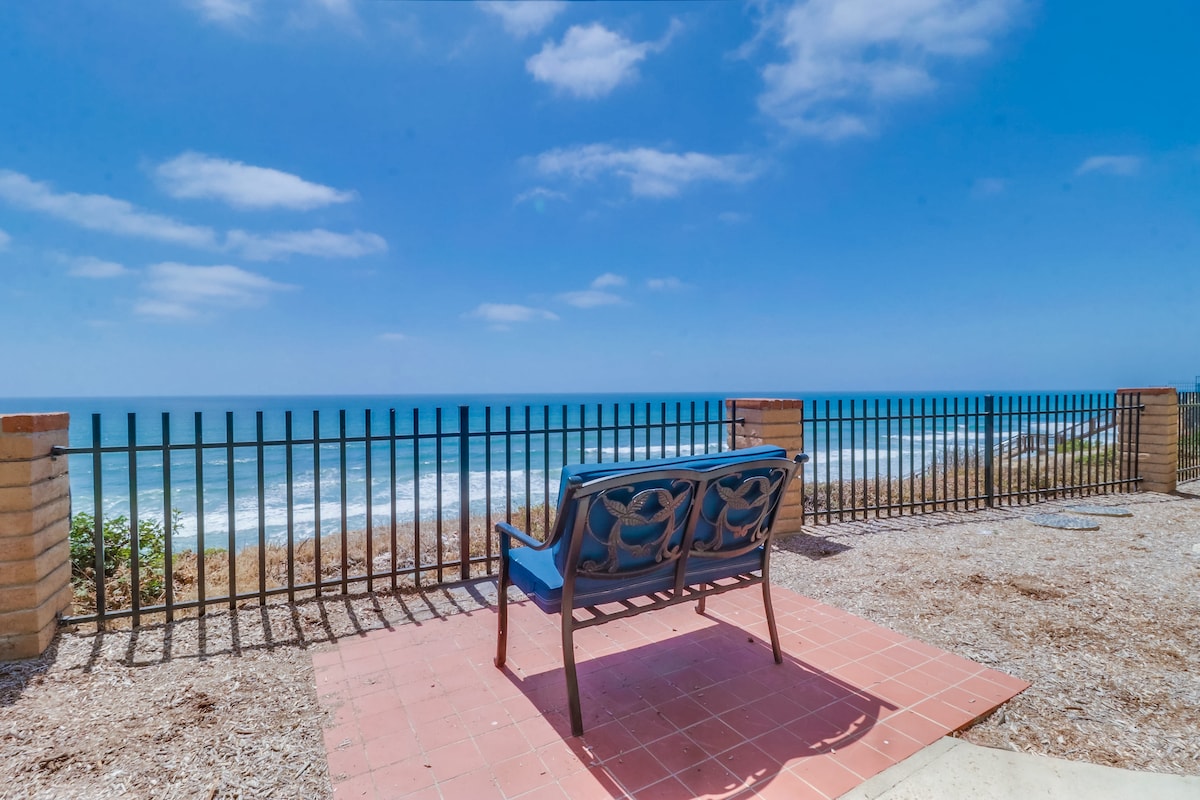 A bench is situated on a tiled patio, overlooking the vast ocean. The scene is framed by a black iron fence, while gentle waves can be seen in the distance under a blue sky with scattered clouds.