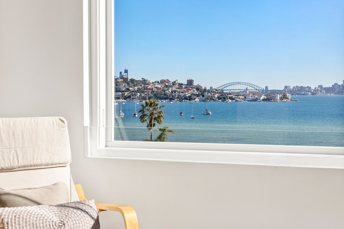 A large window frames a picturesque view of Sydney Harbour, showcasing the iconic Harbour Bridge and sailboats on the water. A palm tree is visible in the foreground, while the bright blue sky complements the serene water.