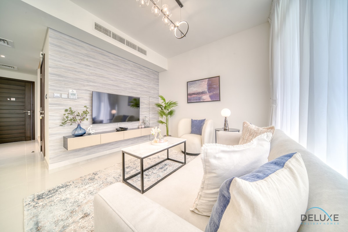 A modern living area is furnished with a comfortable sofa and a glass coffee table. Natural light filters through large windows, while a flat-screen TV is mounted on a textured wall. Decorative plants and artwork enhance the inviting space.