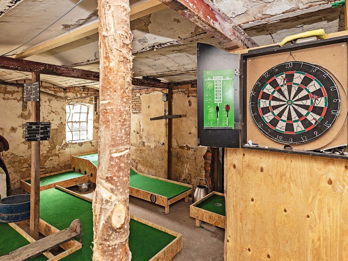 A recreational area features a variety of games, including a dartboard mounted on a wooden panel and two putting greens with vibrant green surfaces. Exposed wooden beams and a rough texture on the walls add a rustic charm to the space.