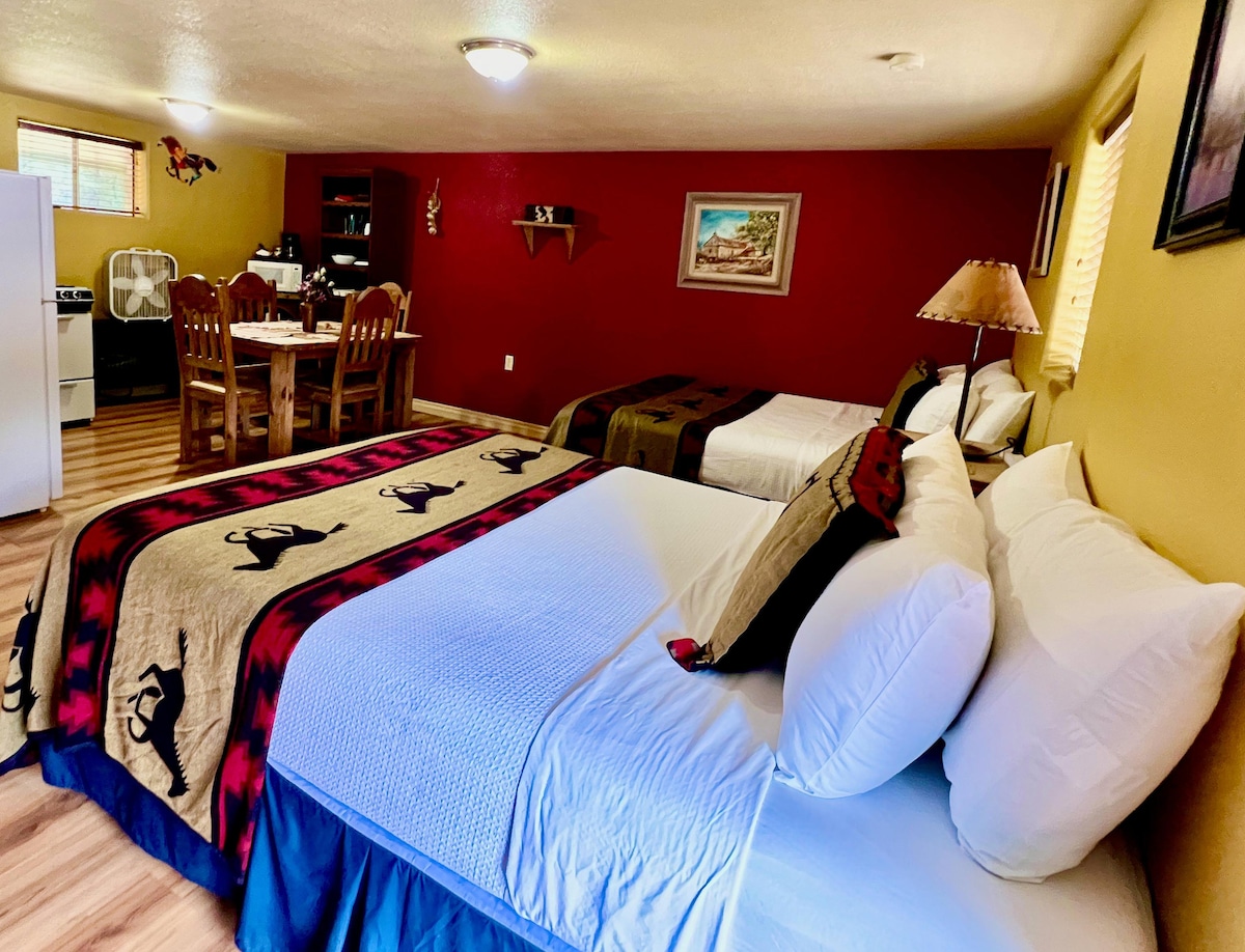 The suite features two Queen beds dressed in inviting linens, accompanied by a cozy throw. A round dining table set for four is visible in the background, showcasing wooden chairs. The warm-toned walls and soft lighting create a relaxed, home-like environment.