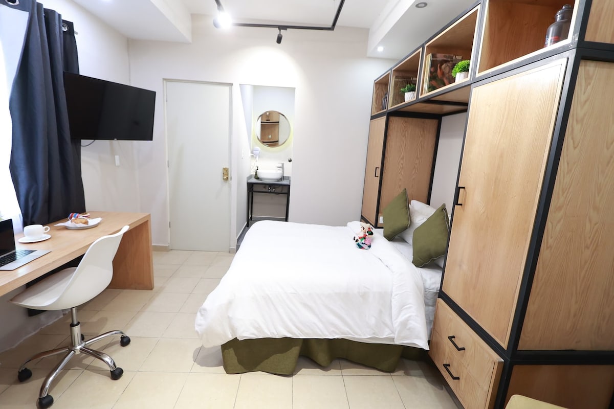 A cozy room is featured, showcasing a spacious bed with green accent pillows. A wall-mounted TV is positioned near a small desk and chair. Light streams through a window covered with blackout curtains, and a compact area for personal items is visible in the cabinetry.