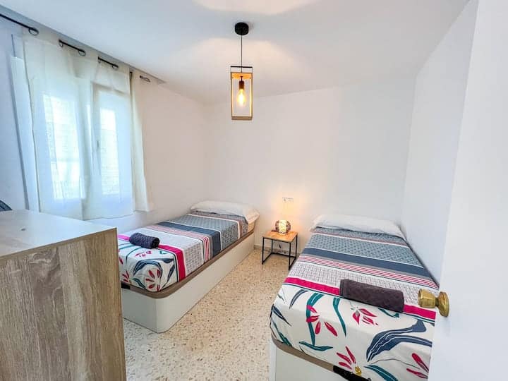 Habitación con dos camas de 90cm. Mesilla de noche con lámpara de sobremesa. Lámpara de techo colgante. Ventana vestida con cortinas.