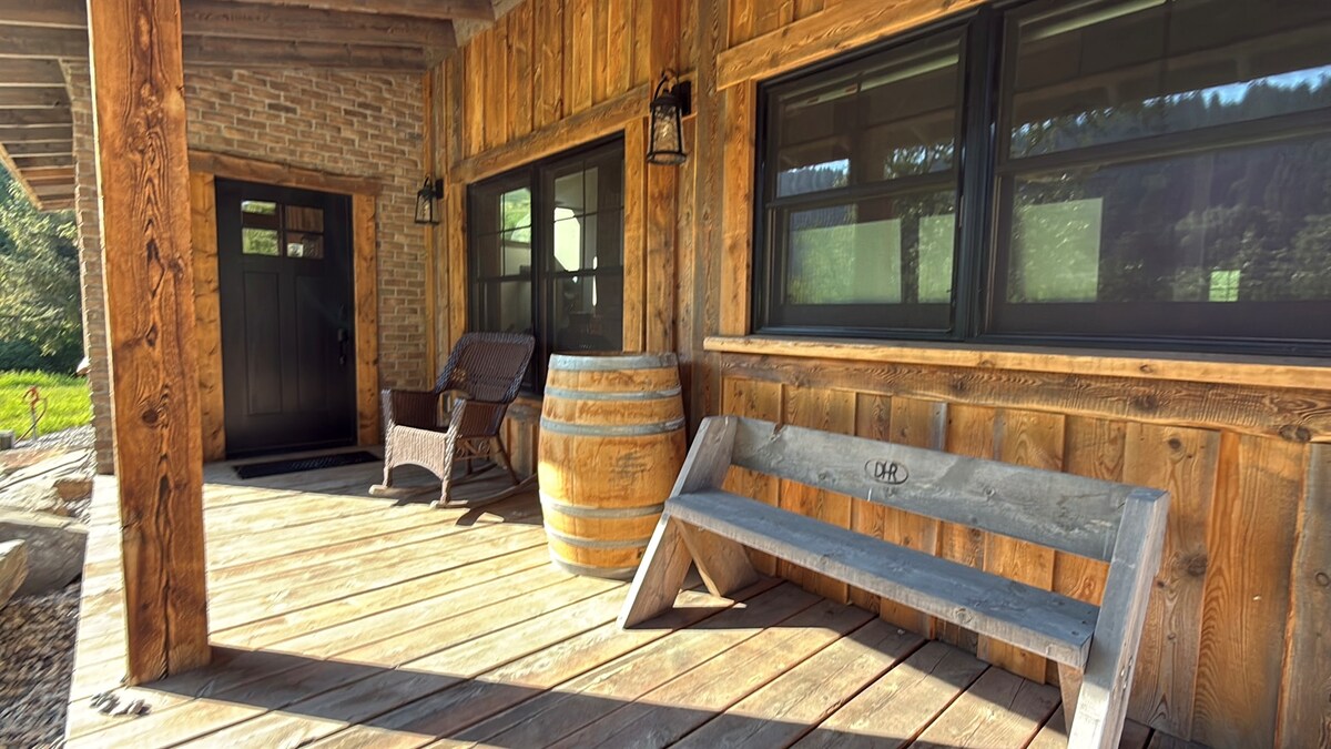 A rustic porch features wooden flooring and walls accented by stone elements. Two lanterns are mounted beside large windows. A wooden bench is positioned beside a wooden barrel, and a comfortable rocking chair adds to the welcoming ambiance.