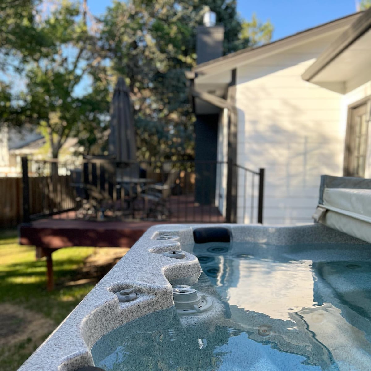 A hot tub is foregrounded, reflecting the clear blue sky. In the background, a wooden deck is visible, shaded by an umbrella. Lush greenery surrounds the space, creating a relaxing outdoor environment.