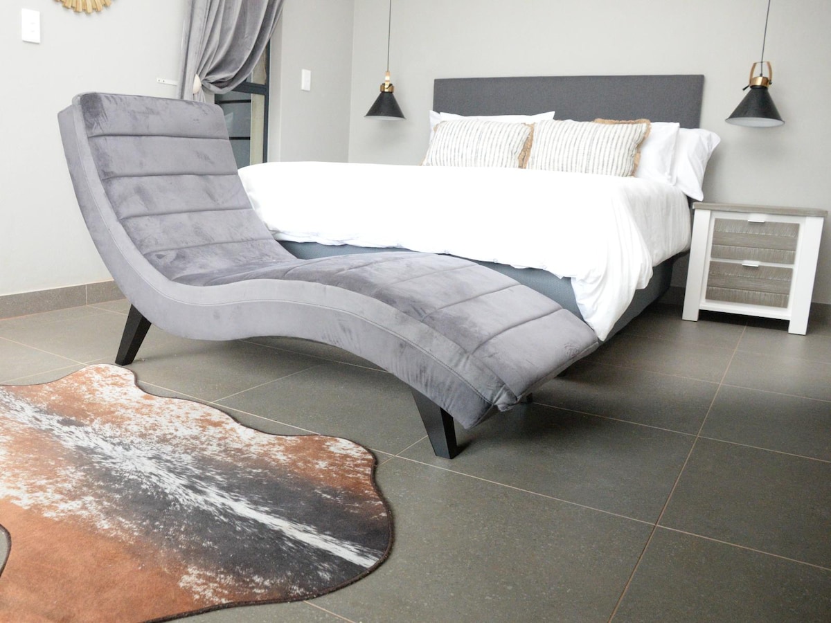 A modern sleeping area features a queen-size bed with crisp white linens and decorative accent pillows. A stylish gray lounge chair complements the space, positioned near a soft area rug. Wall sconces provide subtle lighting on either side of the bed.