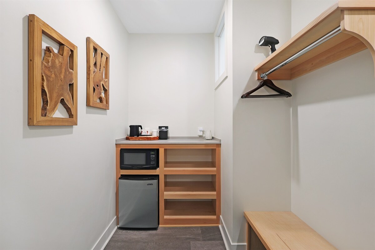 A compact kitchenette area is visible, featuring a refrigerator and microwave. A small shelf and a bench provide space for belongings. Two pieces of wooden wall art add a natural touch to the otherwise simple and functional design.