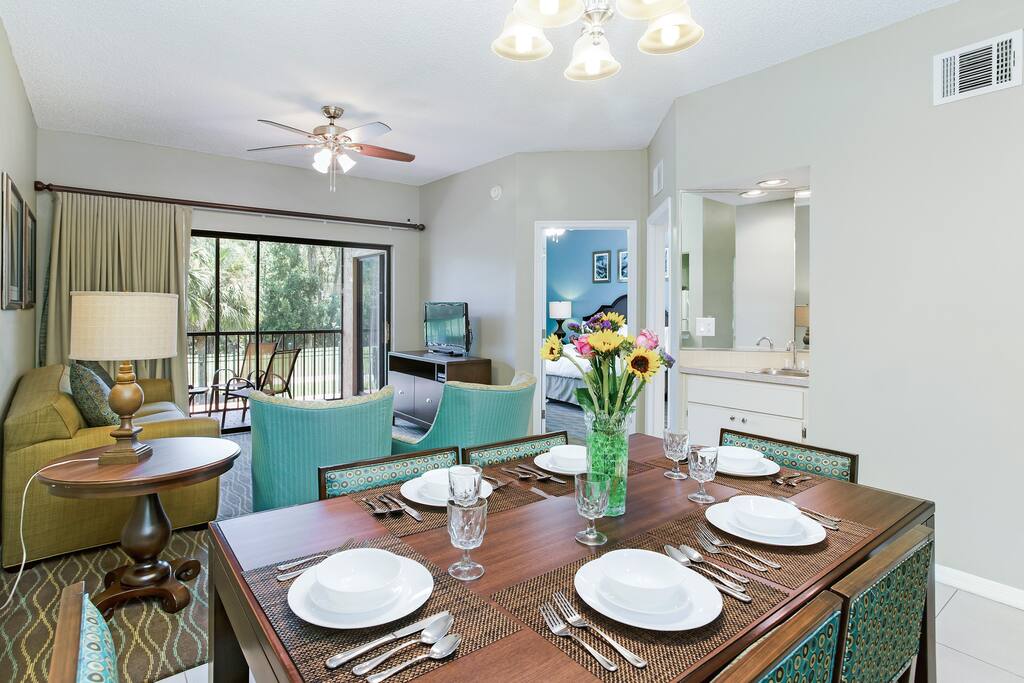 A dining table is set for six, with plates and glasses arranged neatly. In the background, a comfortable sofa and coffee table are seen. A sliding glass door opens to a balcony, offering natural light. Fresh flowers in a vase add a touch of color.