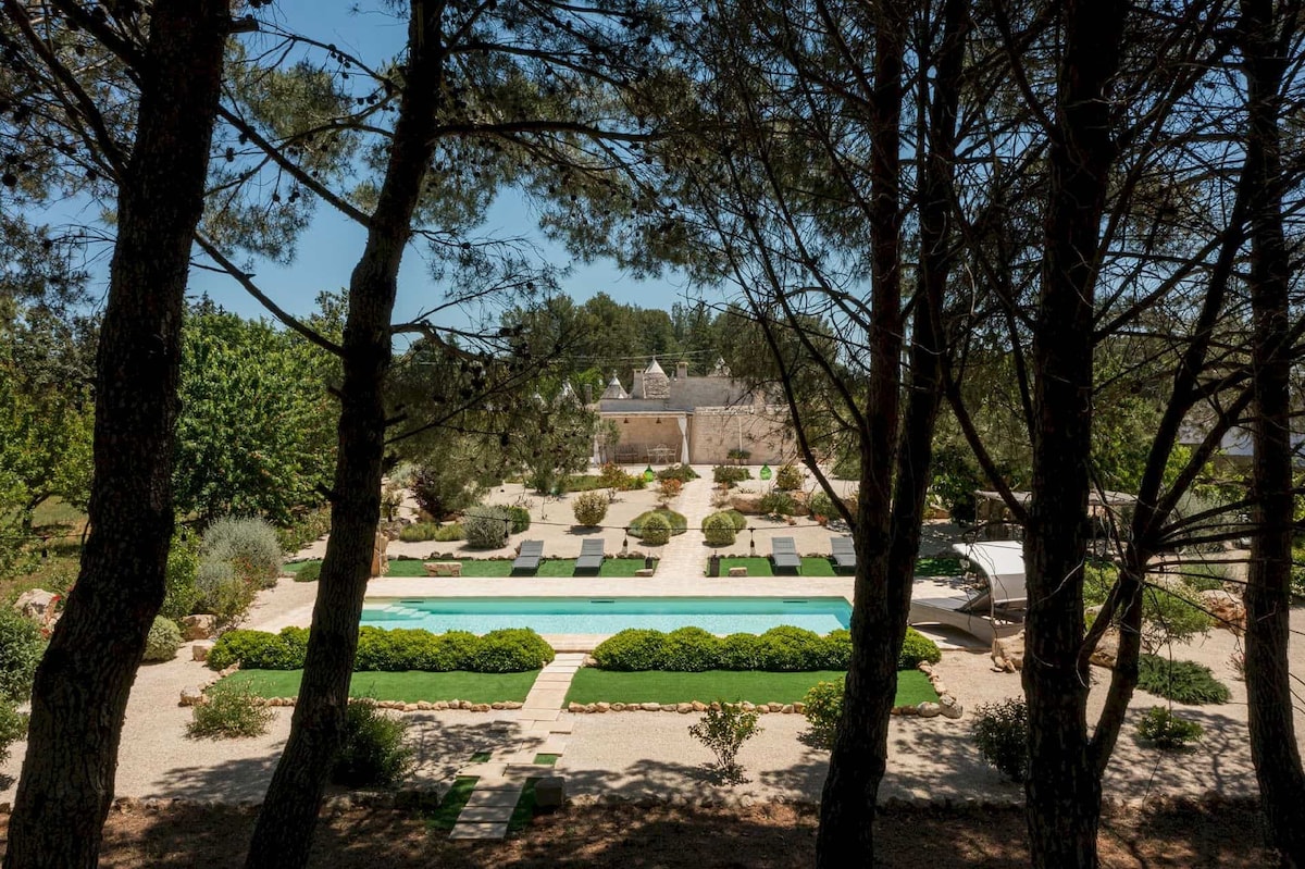 A serene pool area is framed by tall pine trees, showcasing a turquoise swimming pool surrounded by lush greenery and elegant sun loungers. Neatly arranged hedges and garden beds enhance the tranquil atmosphere, creating a peaceful retreat within a beautiful landscape.