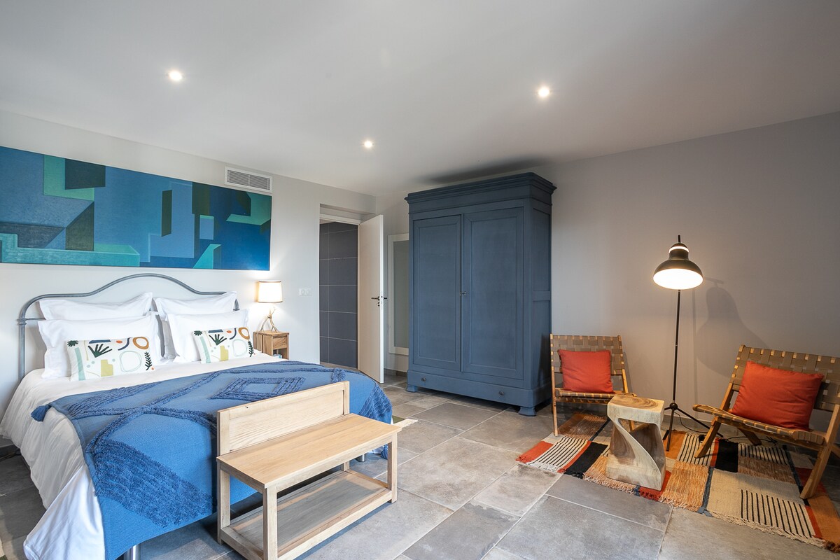 The bedroom features a spacious bed with a blue throw, complemented by an abstract wall art piece. A wooden bench sits at the foot of the bed, accompanied by two patterned chairs with cushions. A lamp and wardrobe complete the inviting space.