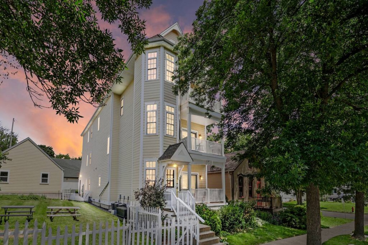 The exterior of a three-story building is framed by lush green trees, with soft evening light glowing from the windows. A white picket fence runs along the front, and a staircase leads to the entrance, enhancing accessibility.