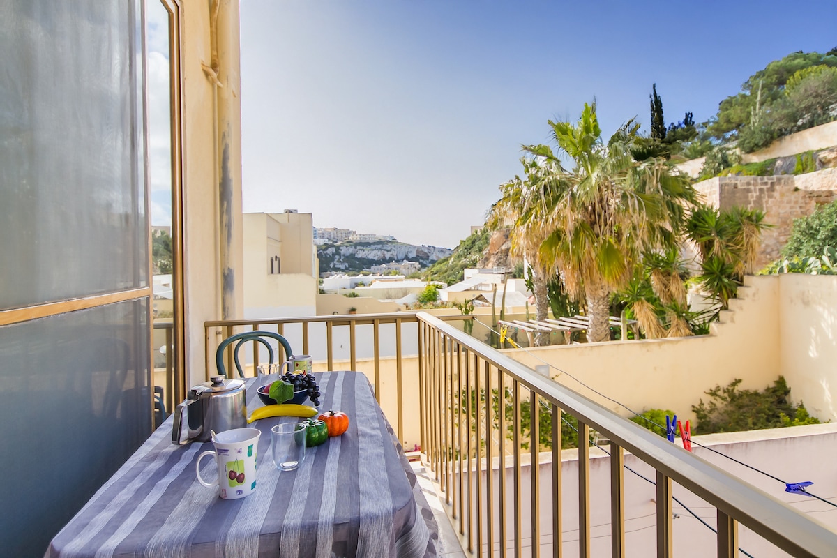 A private balcony is seen with a table set for dining. A kettle, cup, and fresh produce rest on the table, with a view of palm trees and the landscape beyond. Sunlight brightens the space, offering a serene outdoor dining experience.