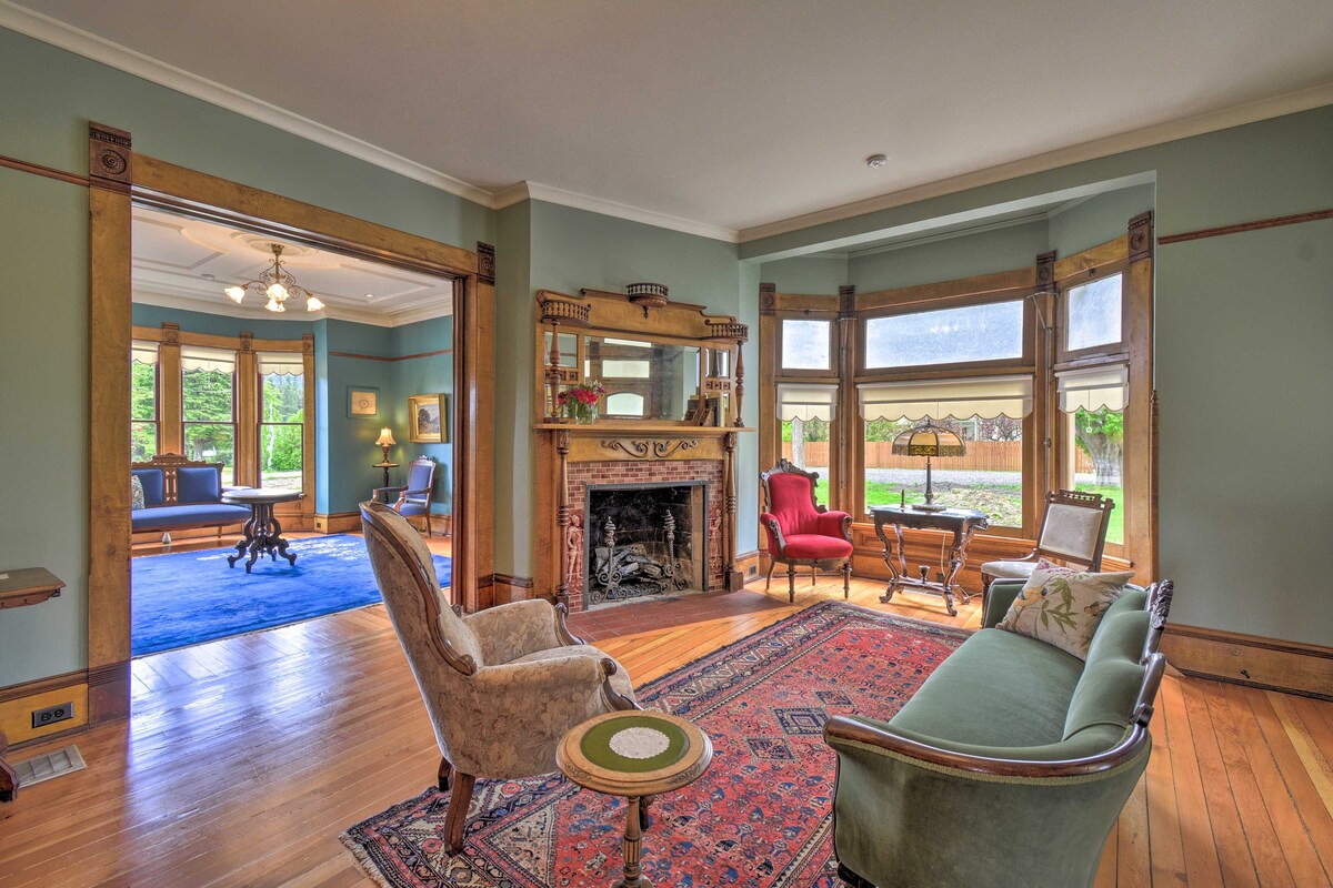 A bright sitting room is highlighted by large windows allowing natural light to fill the space. Victorian-style furniture, including a green sofa and beige armchairs, is arranged around a decorative fireplace. Wooden accents and a colorful area rug enhance the room's historic charm.
