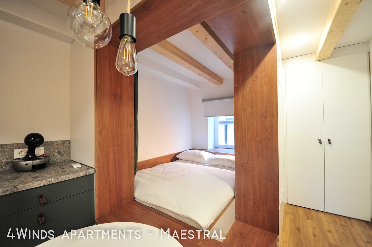 A studio apartment features a double bed integrated within a wooden frame, complemented by exposed ceiling beams. A small kitchenette with a coffee maker is visible in the foreground, while a light-colored closet stands adjacent. Natural light enters through a window, creating a cozy ambiance.