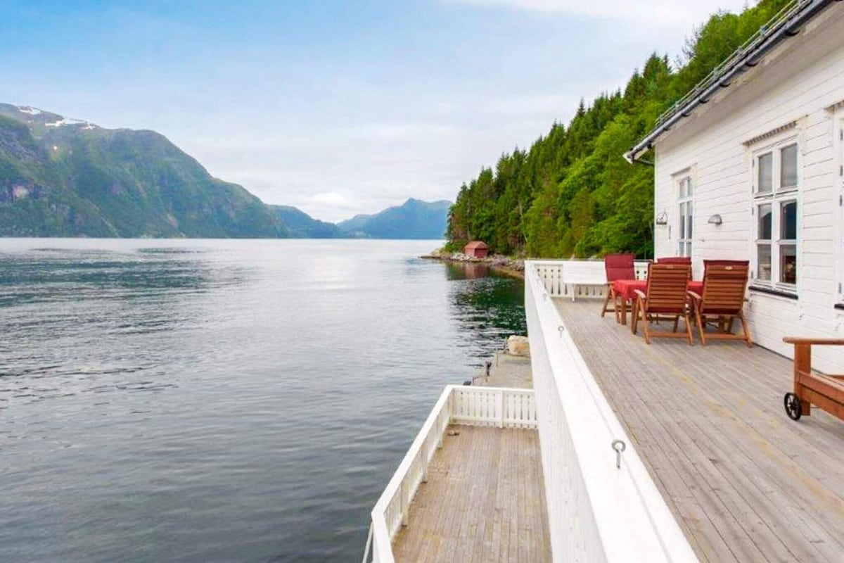 A spacious deck overlooks the calm waters of the fjord, framed by lush green trees and mountainous terrain in the distance. Outdoor seating areas are arranged with wooden furniture, allowing for tranquil moments immersed in nature.