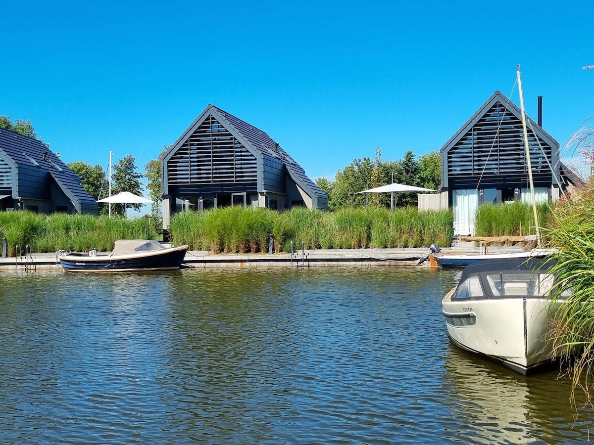 Two modern water villas are positioned along a serene canal, with grassy banks and clear blue skies above. Small boats are anchored at the front, creating a tranquil scene that invites relaxation and exploration.