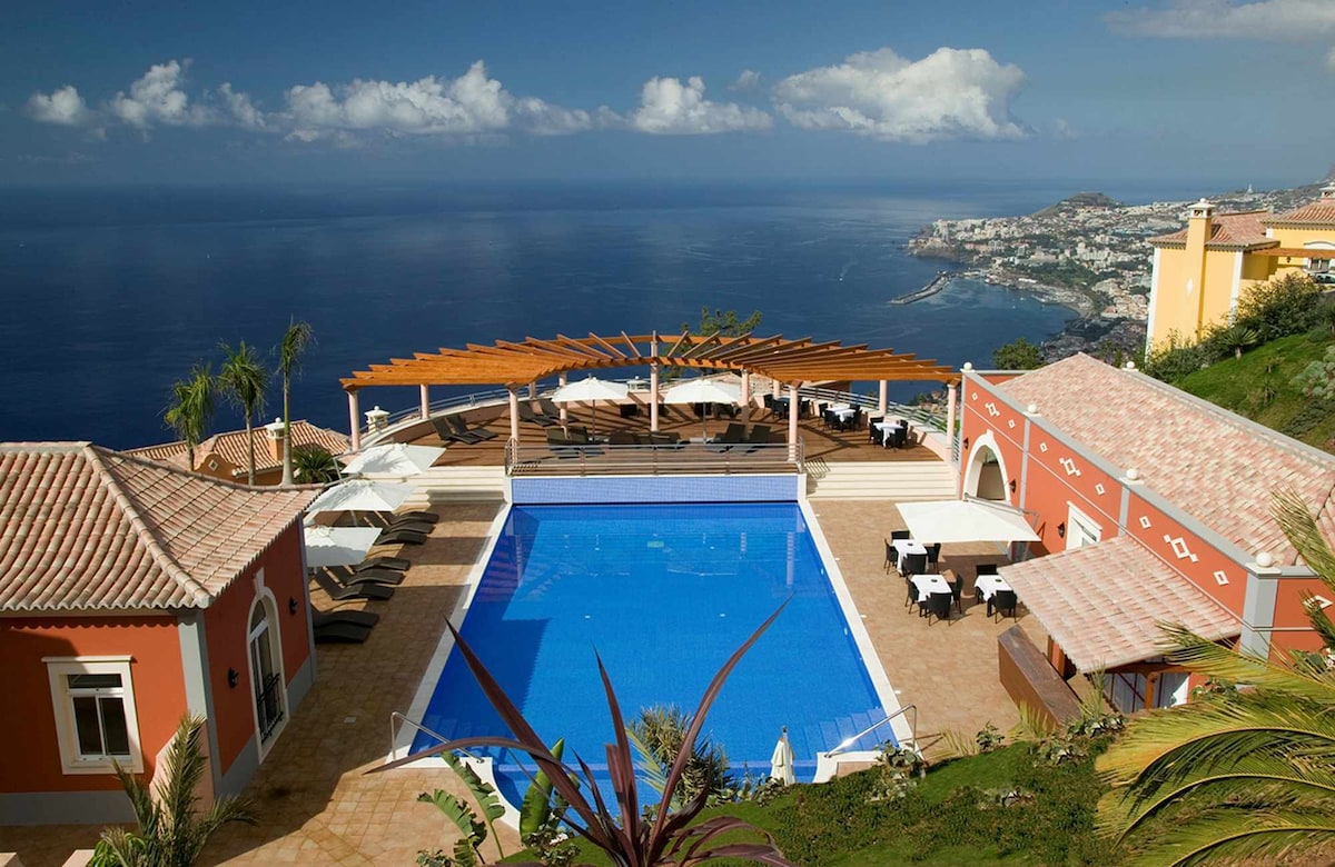 A spacious outdoor area features a large swimming pool with a sun terrace surrounded by lounge chairs. Umbrellas provide shade, and a pergola offers an inviting space for relaxation. The panoramic view includes the coastline and ocean, highlighting the surrounding landscape.