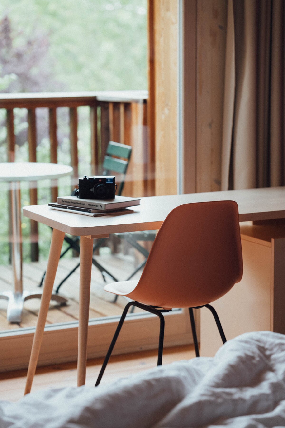 A workspace is positioned adjacent to a large window, allowing natural light to illuminate the area. A simple desk equipped with a chair and a stack of books sits alongside a classic camera. A balcony can be seen through the window, enhancing the serene ambiance.