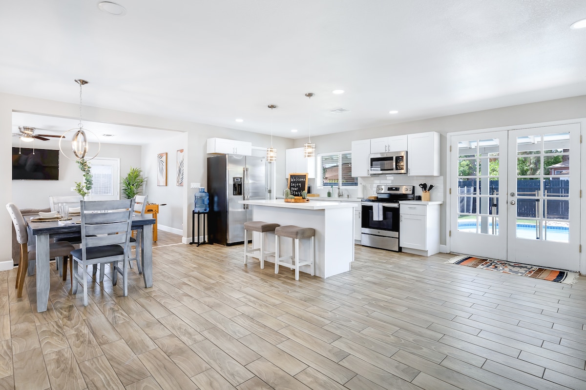 An open kitchen and dining area showcase modern appliances and a spacious layout. The dining table accommodates six, while additional seating at the kitchen island offers casual dining. Large glass doors lead to the outdoor space, providing a view of the pool and backyard.