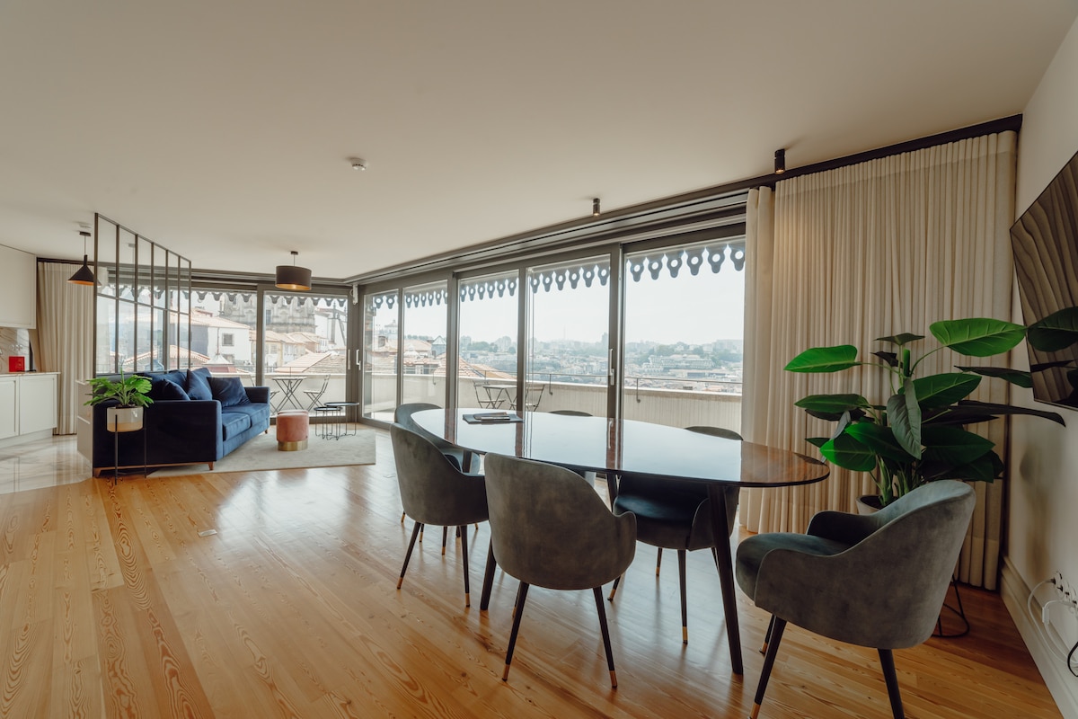 A spacious living area features large glass doors that provide ample natural light and river views. The room includes a modern dining table surrounded by four chairs and a comfortable blue couch. Lush greenery is added by a potted plant in the corner.