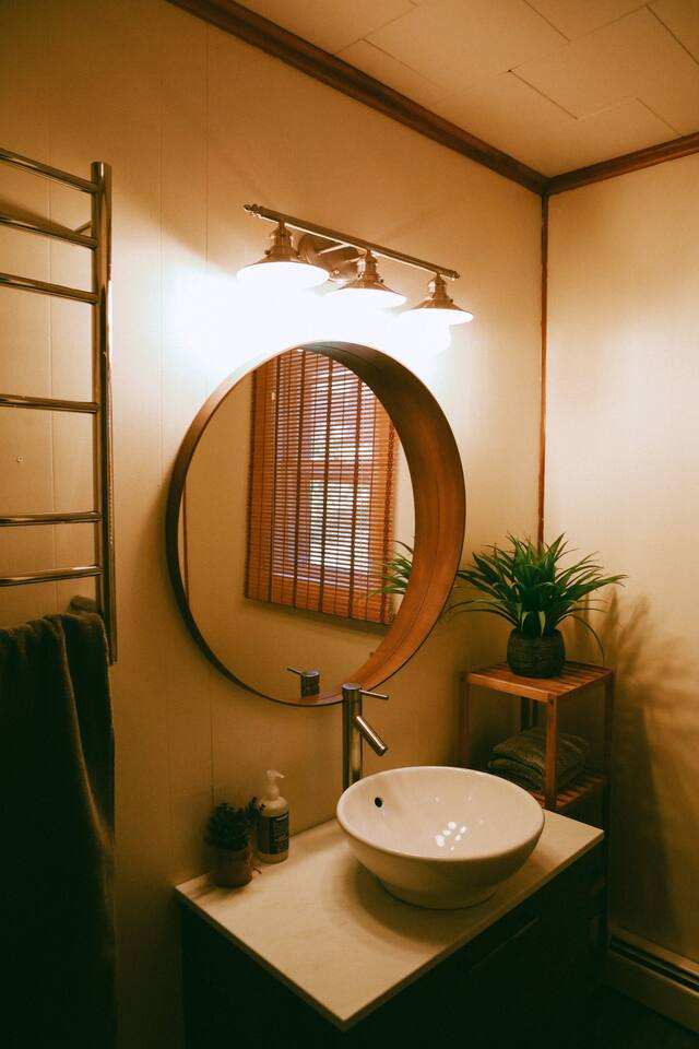 A bathroom features a round mirror illuminated by three light fixtures above. A vessel sink sits on a marble countertop with a modern faucet. Green plants are displayed beside the sink, and a towel rack is mounted on the wall, enhancing the cozy atmosphere.