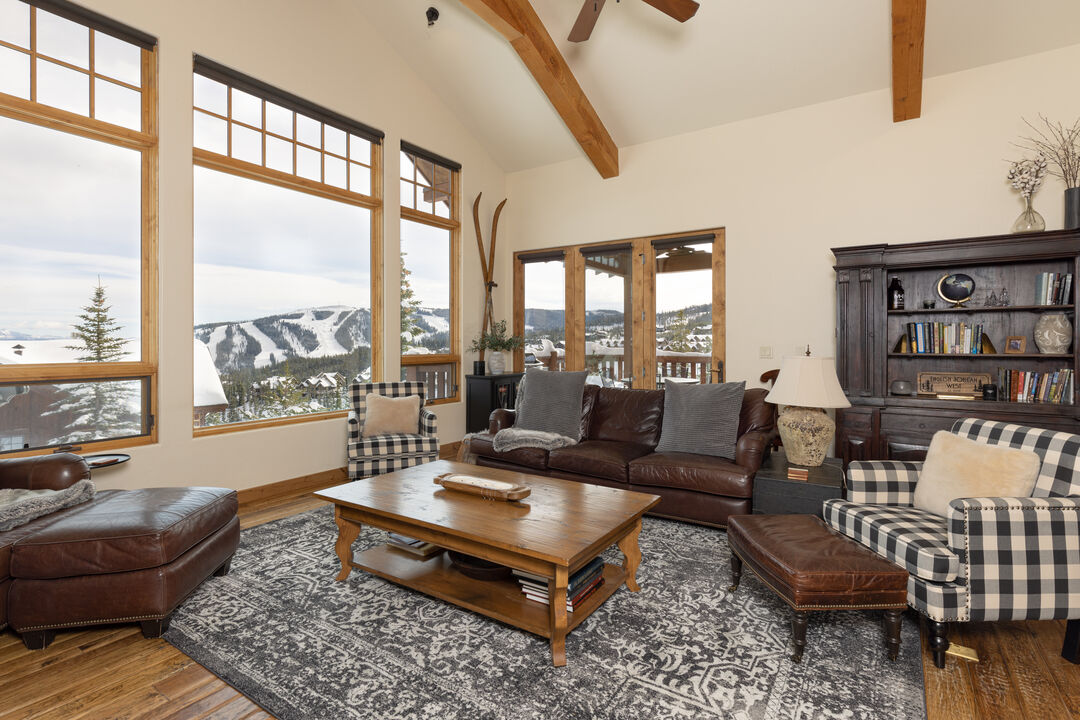 A spacious living area features large windows offering mountain views. Rich leather seating is arranged around a wooden coffee table on a patterned rug. Exposed wooden beams enhance the ceiling's height, while a bookshelf and decorative elements add warmth to the space.