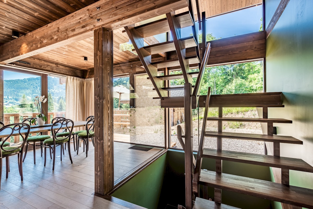 An open-concept living space features large windows allowing natural light to fill the room. A stylish dining area is positioned near the windows, accompanied by wooden stairs that lead to an upper level. The design incorporates natural materials, blending seamlessly with the surrounding landscape.