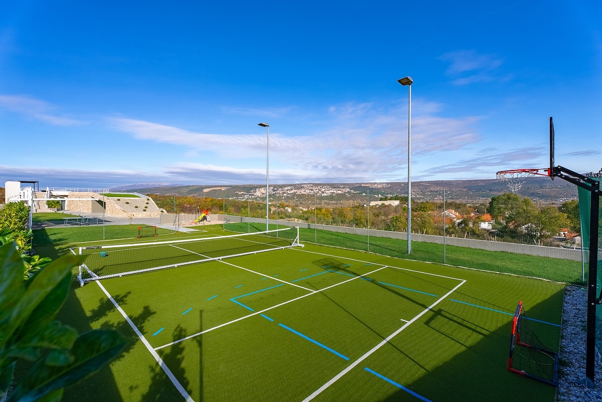 A multi-sport play area features a well-maintained artificial grass surface, marked for mini soccer, tennis, and basketball. Surrounding the court is a protective fence and open views of the countryside, while bright lighting fixtures are positioned for evening use.