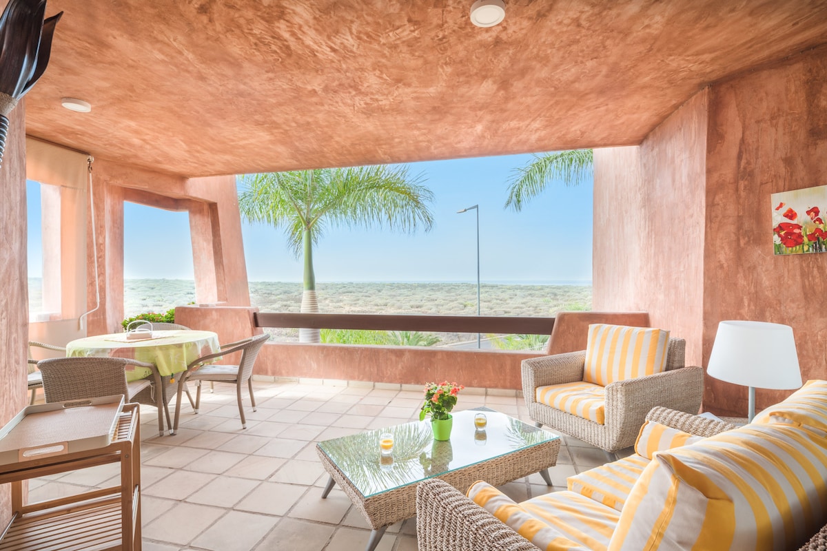 A covered terrace features comfortable seating with beige and white striped cushions. A small glass table sits between the chairs, complemented by a potted plant. Large windows showcase panoramic views of the sea, enhanced by palm trees in the background.