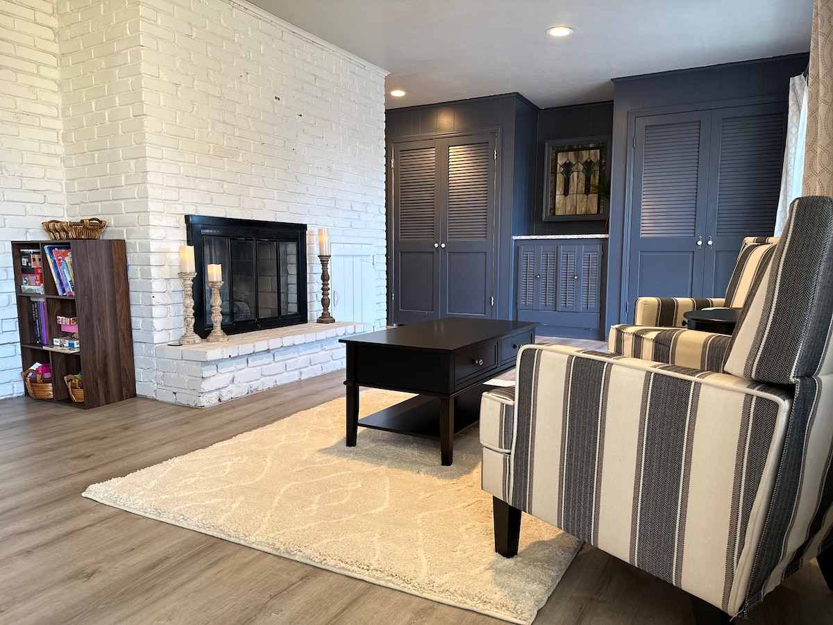 A cozy living area features a white brick fireplace as a focal point. Two striped armchairs are positioned beside a dark coffee table on a soft rug. A wooden bookshelf displays various items, while cabinetry and a wall art piece add character to the space.