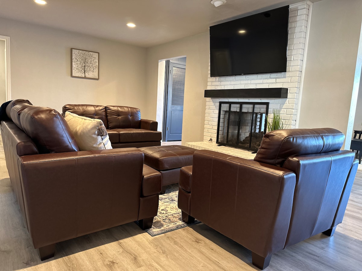 A welcoming living area is arranged with brown leather seating, including two sofas and two armchairs, placed around a coffee table. A modern fireplace with white brick accents serves as a focal point, while a large flat-screen TV is mounted above the mantel, enhancing the space.