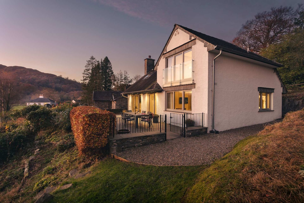 The exterior of a modern home is shown at dusk, featuring large windows that reflect the soft evening light. A spacious deck is visible, accompanied by outdoor seating. Surrounding greenery and hillside views add to the appeal of the serene location.