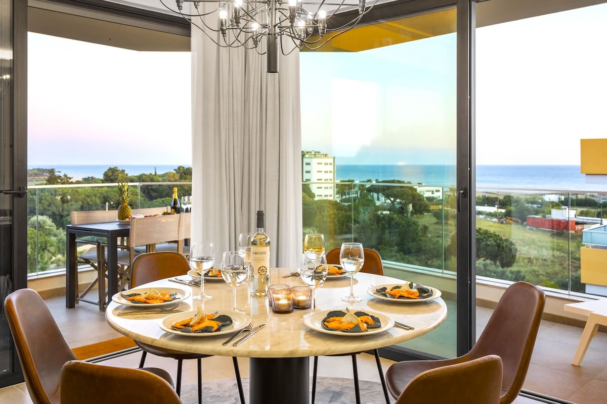A dining area features a round table set for four, complete with plates, glasses, and a bottle of wine. Floor-to-ceiling windows frame ocean views and bring in natural light. A stylish chandelier hangs above the table, enhancing the modern decor.
