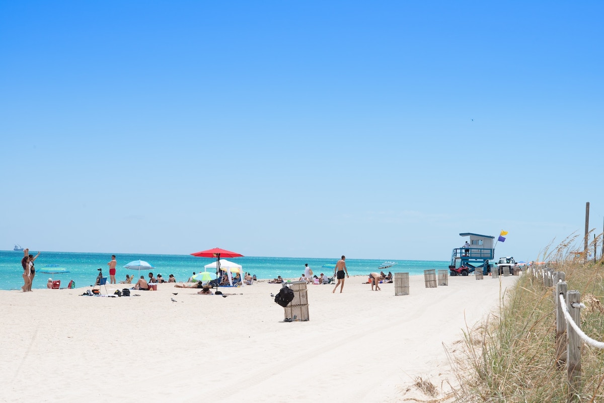 A sandy beach area is visible, with various umbrellas providing shade for sunbathers. A lifeguard tower stands nearby, overseeing the clear blue water. A pathway lined with wooden frames leads toward the shore, where beachgoers relax under the warm sun.
