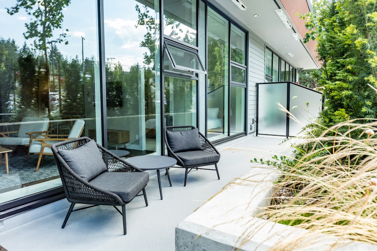 An outdoor private deck features two contemporary lounge chairs and a small side table, surrounded by greenery. Large windows provide a view of the lush surroundings, contributing to a relaxed outdoor setting.