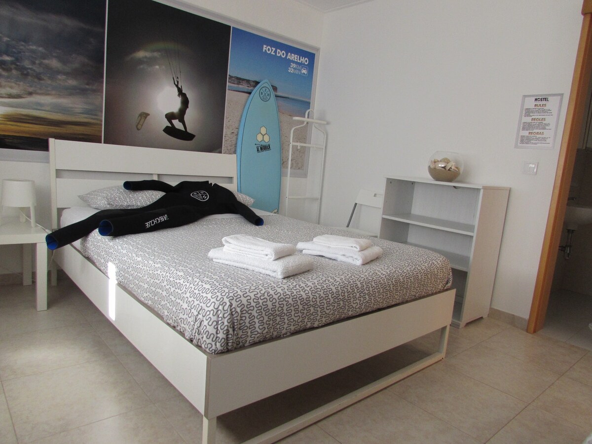 A spacious bedroom is presented with a white bedframe and patterned bedding. Several folded towels are arranged neatly atop the bed. Surfing equipment, including a wetsuit and surfboard, enhances the room's coastal theme. A white dresser and minimal decor contribute to the airy atmosphere.