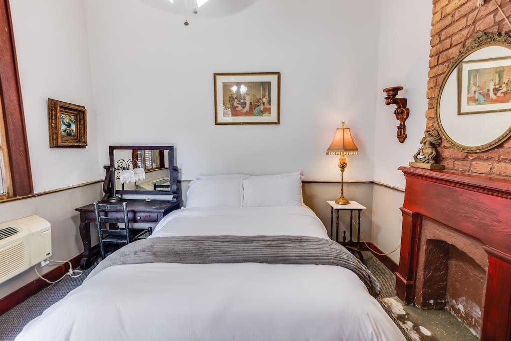 A comfortable bedroom features a spacious bed dressed in crisp white linens. A small bedside lamp provides soft light beside a vintage-style mirror and dresser. Decorated with framed art, the space includes a charming brick fireplace against the wall.