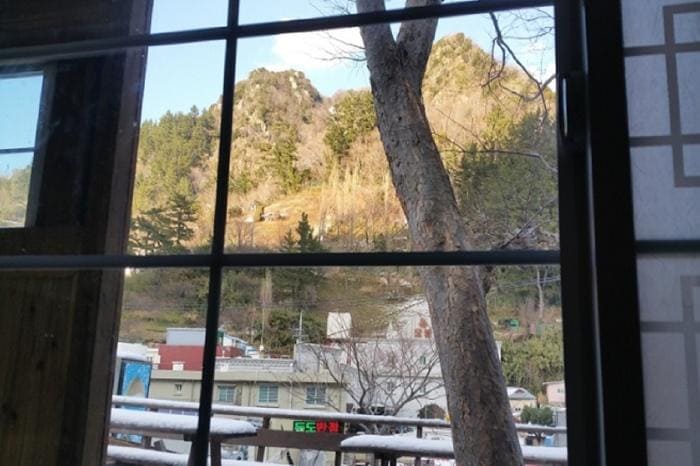 A view from a window showcases a mountainous landscape, with trees lining the foreground. Buildings are visible below, with a hint of colorful signage. Natural light enhances the scene, highlighting the textures of the mountain and the urban surroundings.