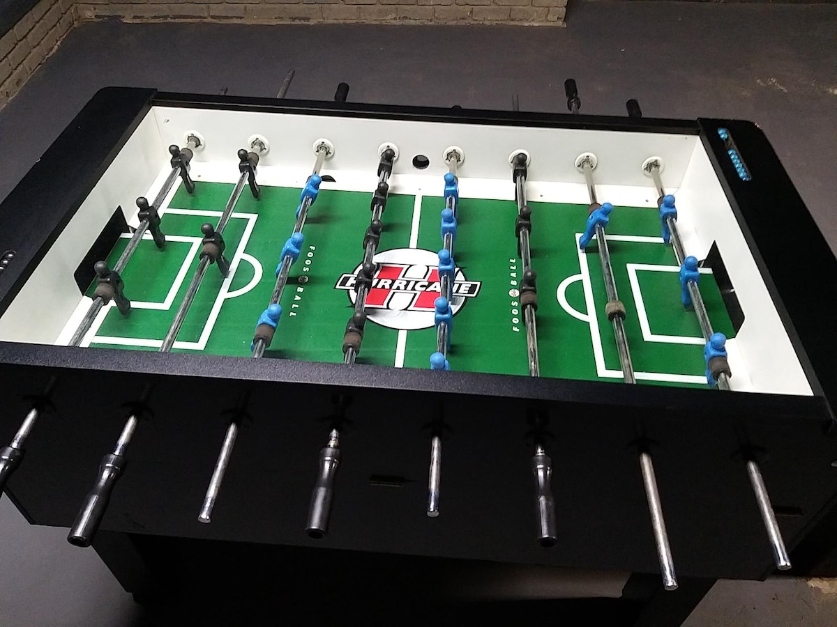 A foosball table with a green field is positioned in an indoor space. The table features black and blue playing rods, with several figures attached for gameplay. The logo of 'American' is prominently displayed at the center, surrounded by goals on each end.