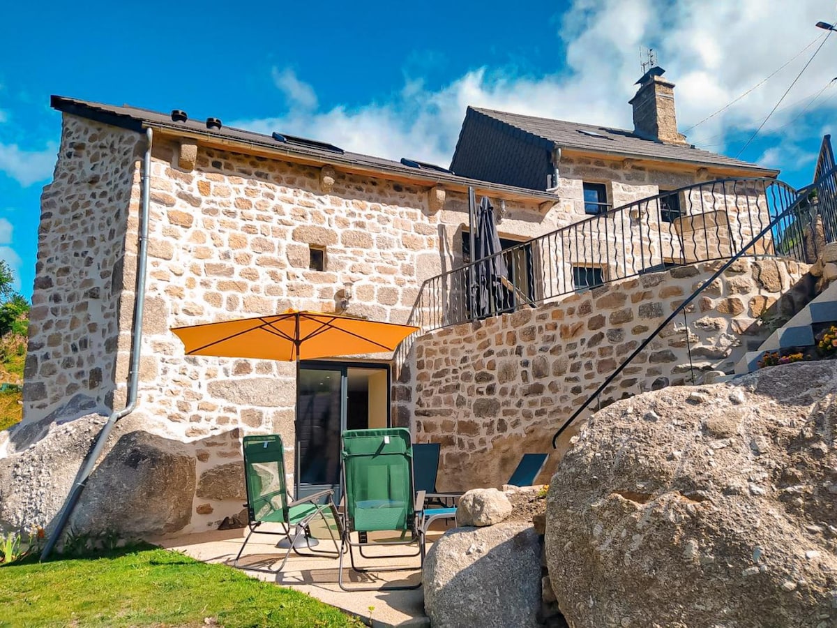 The exterior of the gîte features natural stone walls and a patio area with two green loungers beneath an orange umbrella. Stairs lead to an upper level, while large glass doors provide access to the interior. The surrounding landscape includes prominent granite boulders.