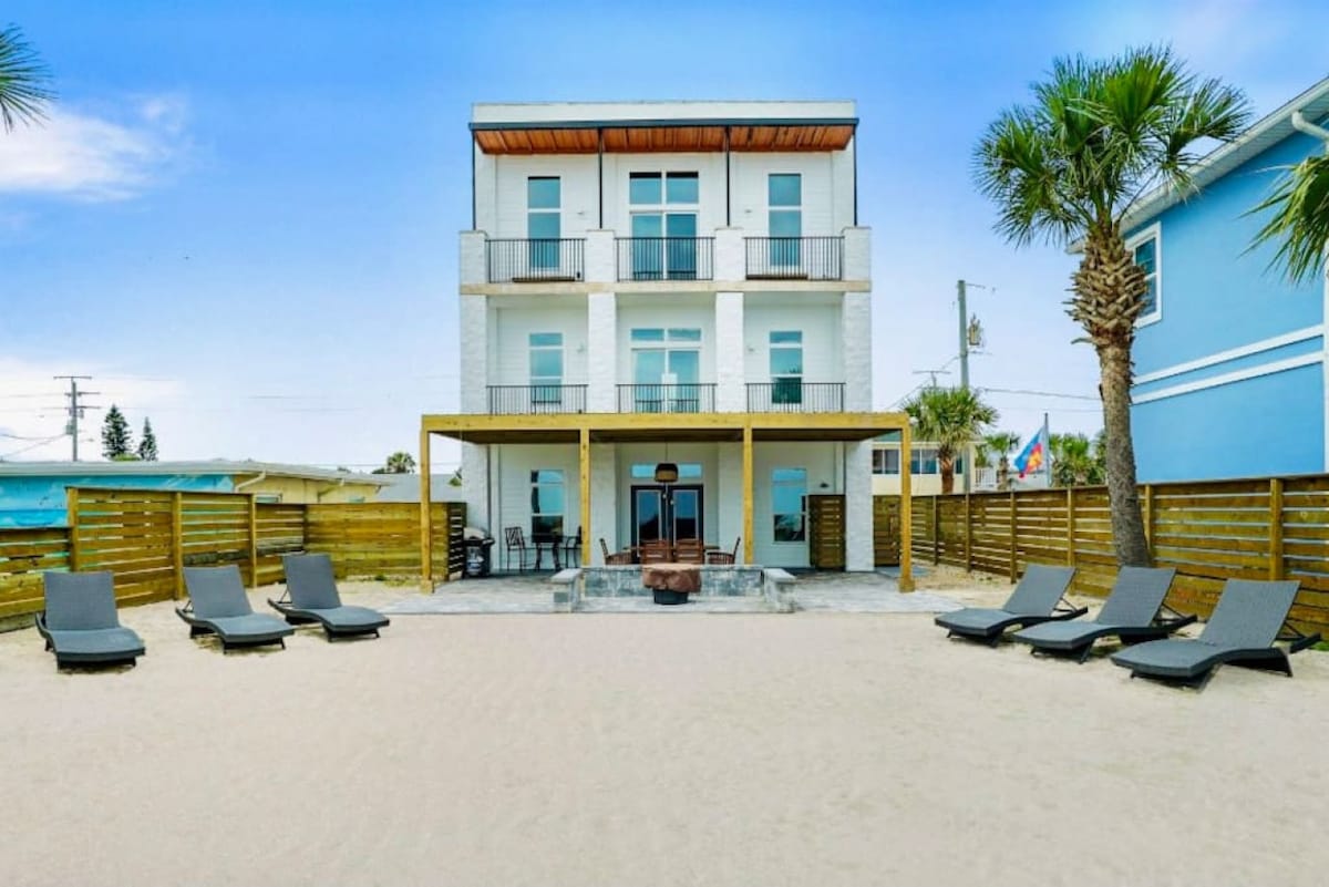 A three-story beachfront home features a covered outdoor area with a fire pit and ample lounging space. Several loungers are positioned on the sandy beach, offering a view of the house's modern architecture and palm trees that surround the property.