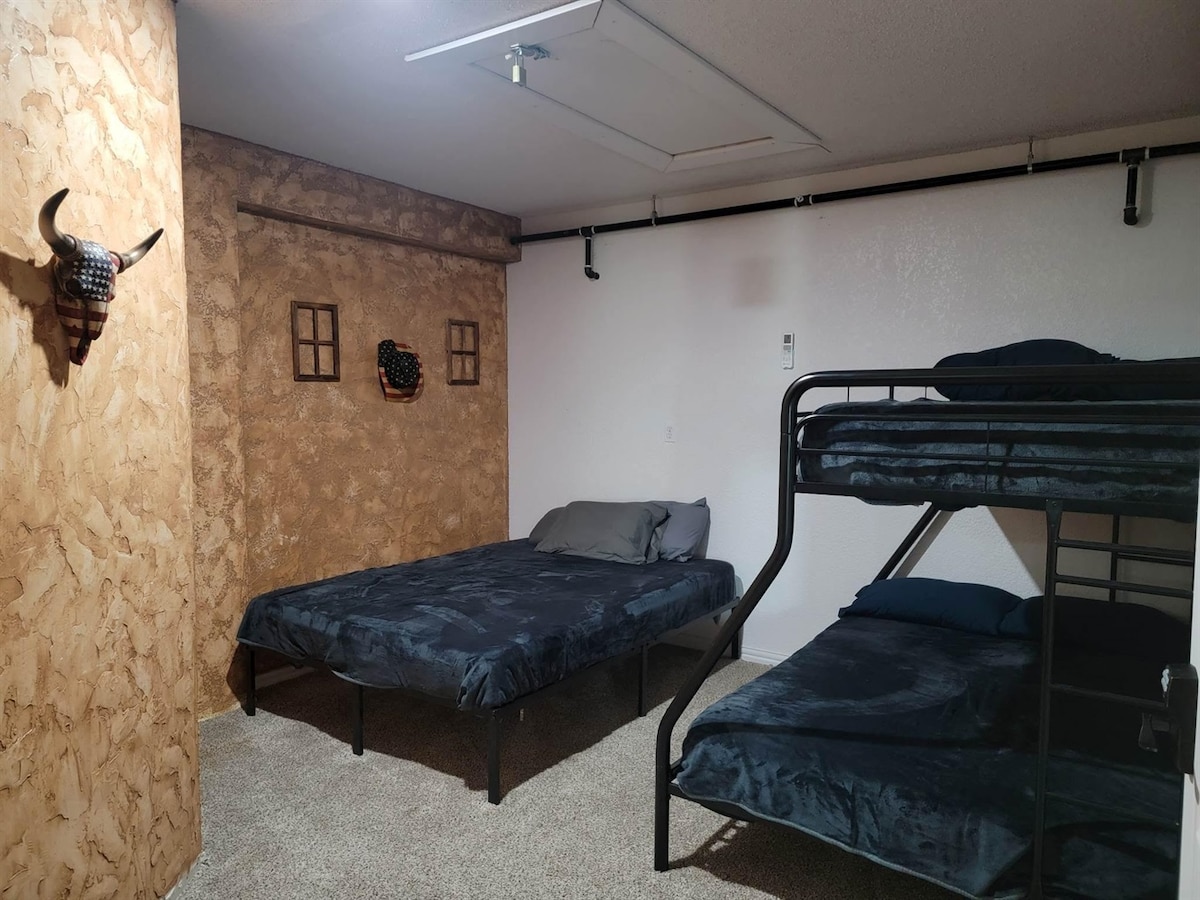 The sleeping area features a spacious bed alongside a bunk bed. Textured walls with rustic accents are present, and two small windows contribute to the natural light. A neutral carpet covers the floor, enhancing the cozy environment.