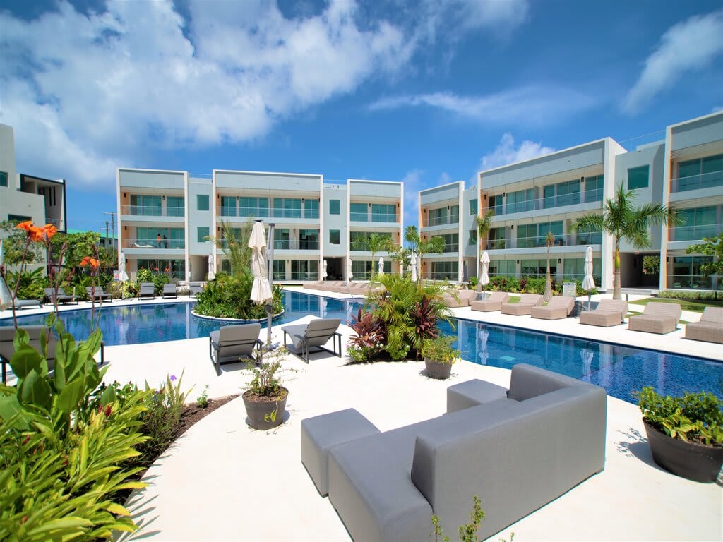 The outdoor area showcases a modern pool surrounded by lush greenery and lounge chairs. A sleek, elongated seating arrangement is positioned nearby, offering a relaxing space to enjoy the sun. The building’s contemporary design features large windows and balconies that enhance the serene atmosphere.
